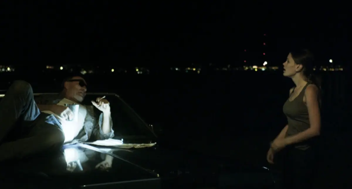 At night, a man in sunglasses lounges on the hood of a car under a harsh light, talking to a woman standing in the dark, with city lights across the water behind them.