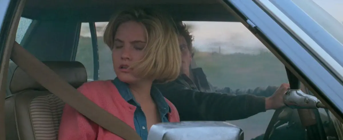 A blonde woman in a pink cardigan sits with her eyes half closed in a car’s front seat while a dark-haired man reaches past her for the steering wheel.