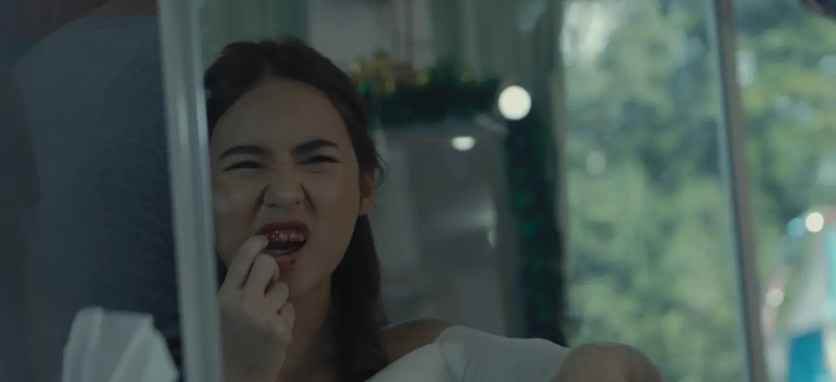 A young woman grimaces into a mirror, her teeth and lips stained with blood as she touches her mouth with her fingers.