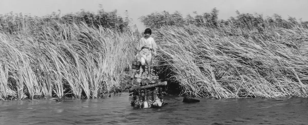 Black-and-white still: a young woman carrying a wooden bucket steps out of a narrow gap in tall, wind-bent reeds toward a river’s edge, where a bundle of armour and cloth lies half-submerged in the water.