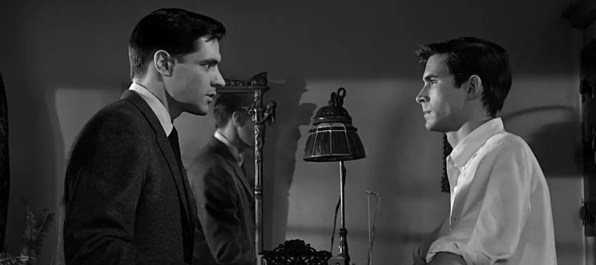 Black-and-white still: a young man in a dark suit faces Norman Bates across a dim parlour, an old lamp between them and the young man’s reflection in a mirror behind.