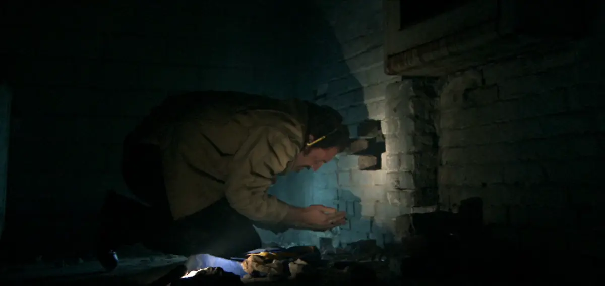 In a dark basement, a man in a work jacket kneels by a flashlight and peers into his cupped hands beside a hole in a crumbling brick wall.