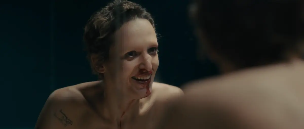 A bare-shouldered woman with short, slicked-back hair and a bloodied nose and mouth grins at a blurred figure in the foreground.